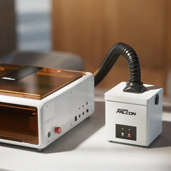 Echo Six Hobbies Creality Falcon smoke purifier with a black flexible hose connected, placed beside a white enclosed laser engraver on a desk