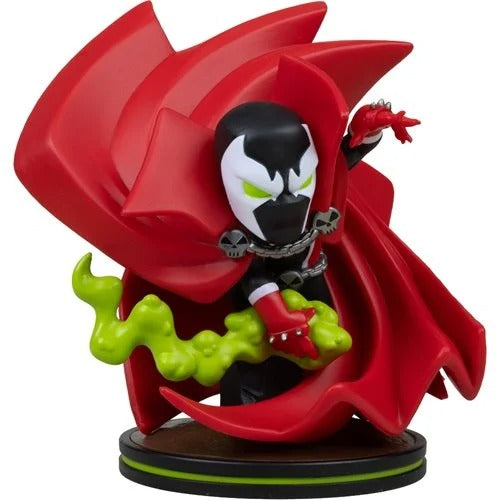 A stylized collectible figure of Spawn with a large flowing red cape, black and white mask, green energy effects, and a round display base on a plain white background.