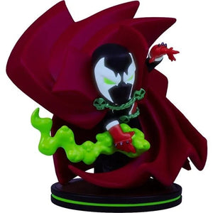 A small stylized figurine of a hooded character in a large dark red cape, with a black face, white mask area, green glowing eyes, red gloves, and a bright green slime-like effect, standing on a black circular base against a plain white background.