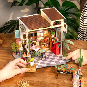 Miniature model of a cafe with plants and a checkered floor, held by hands on a wooden surface.