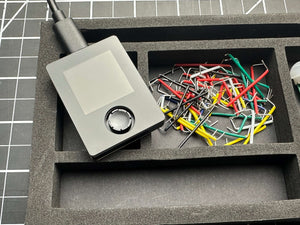 Compact electronic device with a display and control dial alongside assorted colored wires in a foam tray, supporting STEM learning with Make: Getting Started with Oxocard Connect - PDF, from a veteran-owned hobby store.