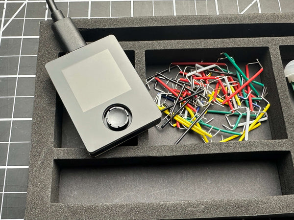 Compact electronic device with a display and control dial alongside assorted colored wires in a foam tray, supporting STEM learning with Make: Getting Started with Oxocard Connect - PDF, from a veteran-owned hobby store.